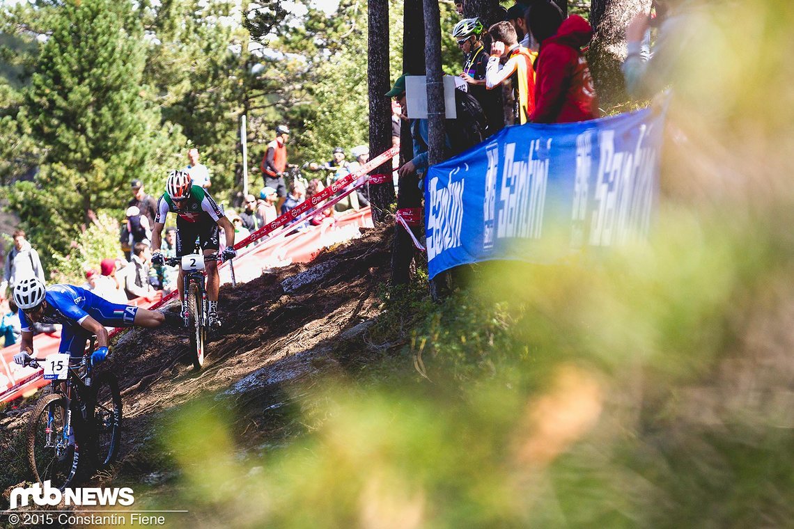 Andrea Tiberi bekommt Probleme sich in der ersten Runde auf dem Rad zu halten, während Schurter elegant über die nassen Wurzeln fährt.